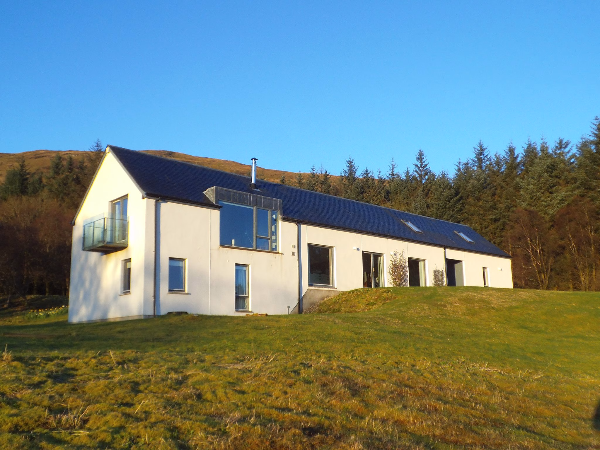 The bright white, modern Corry House with large windows and a dark roof, situated on a green hillside and backed by a dense forest.