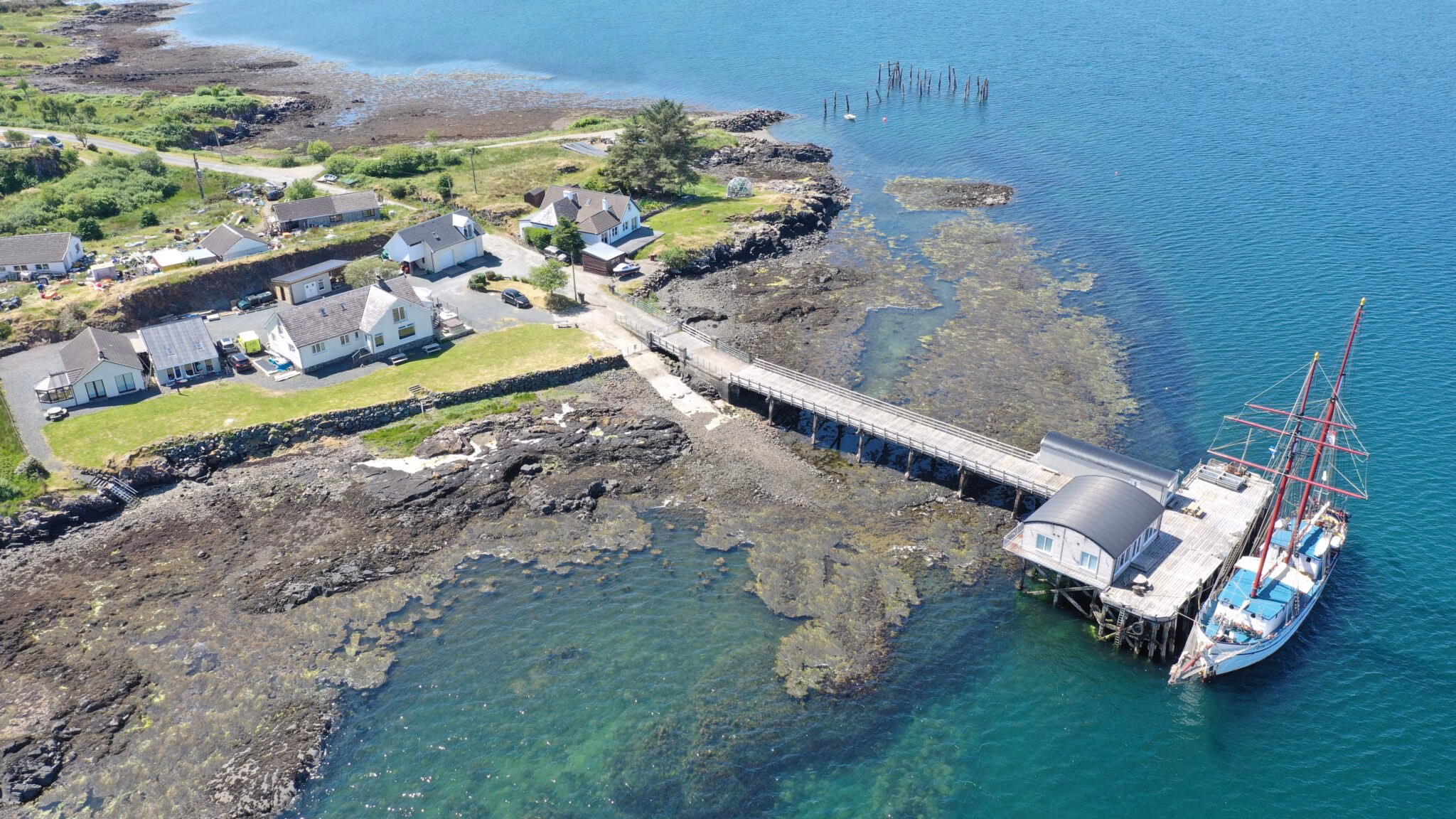 Aerial view of the Salen Pier Holidays complex, showing white cottages on the shore, a long wooden pier extending into the turquoise water, and a large sailboat moored at the end.