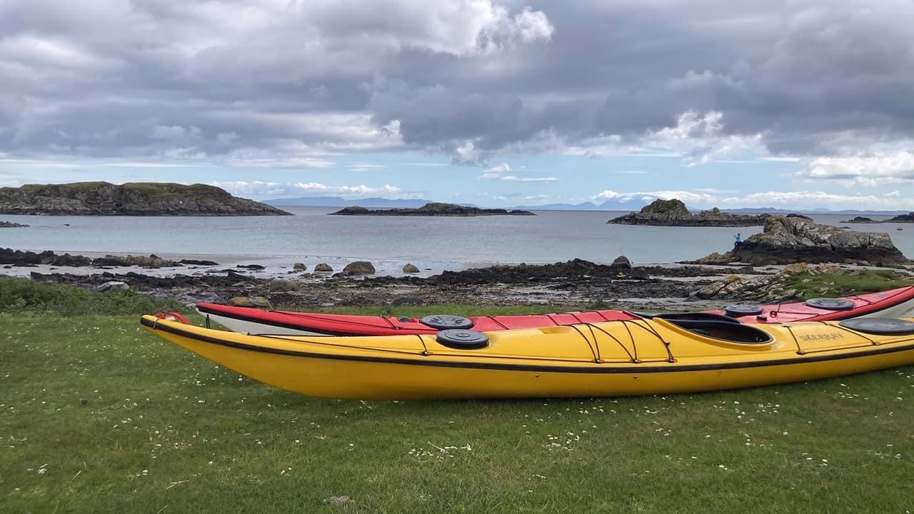 A yellow sea kayak and a red kayak rest on the grass in the foreground of a beach. Rocky islands are visible across the calm water under a cloudy sky.