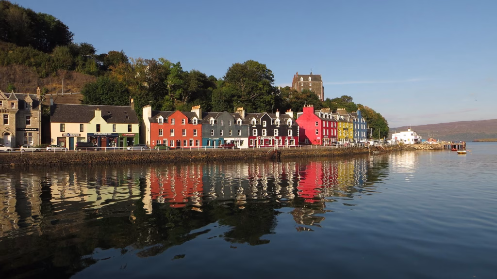 The iconic brightly colored houses of Tobermory line the harbor wall, with their vivid reflections visible in the calm water, and a dark tower house on the hillside above.