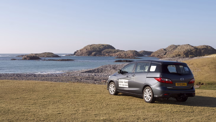 The gray Iona Taxi minivan is parked on a grassy verge overlooking a rocky beach, with the company's logo visible on the side and granite islands in the background.